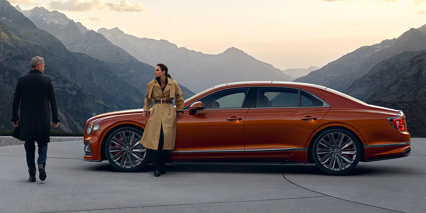 Bentley Suomi Bentley Flying Spur Speed parked in Orange Flame coloured exterior parked, with mountainous background and two people in view.