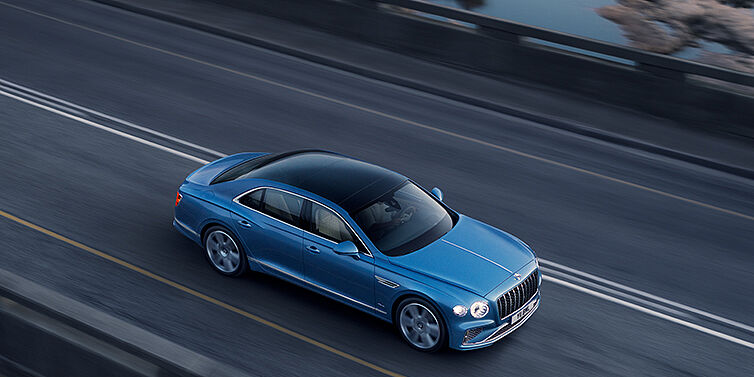 Bentley Suomi New Bentley Flying Spur Azure sedan in Light windsor blue paint by Mulliner, with 22 inch Azure wheels seen from overhead driving dynamically on a country road