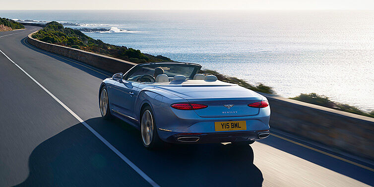Bentley Suomi New Bentley Continental GTC Azure convertible rear view in Light Windsor blue paint with 22 inch ten spoke wheels driving dynamically on an ocean road in the sunshine